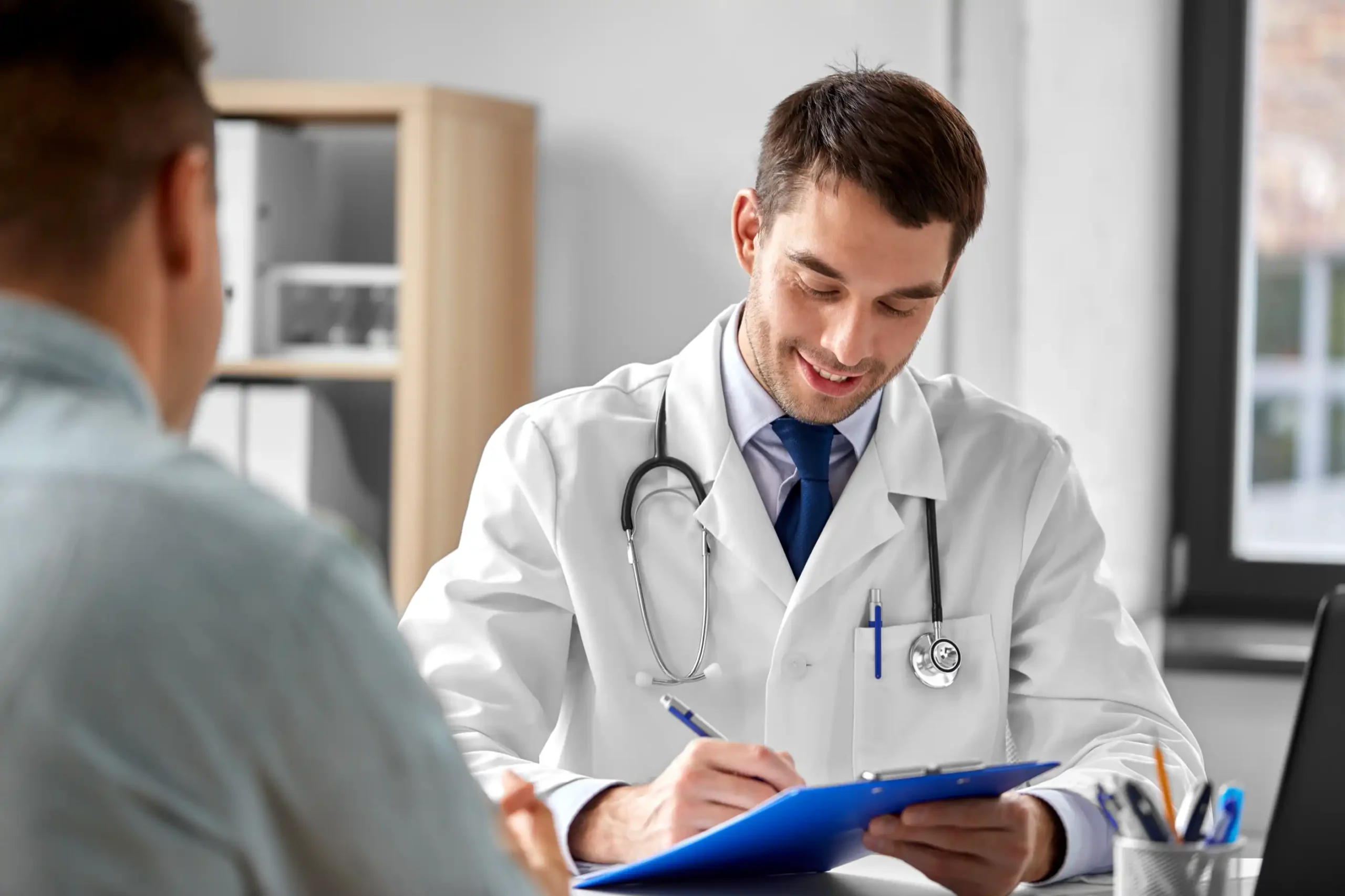 Smiling injury doctor taking notes during a patient consultation, representing personalized care at your first visit with ImpactMD’s treatment team.