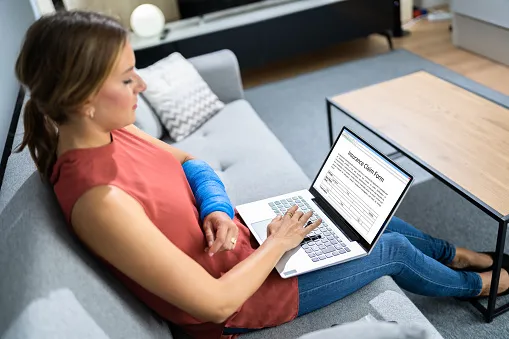 Woman with her arm in a cast typing on a laptop at home, highlighting the financial concerns and recovery needs after a car accident injury.