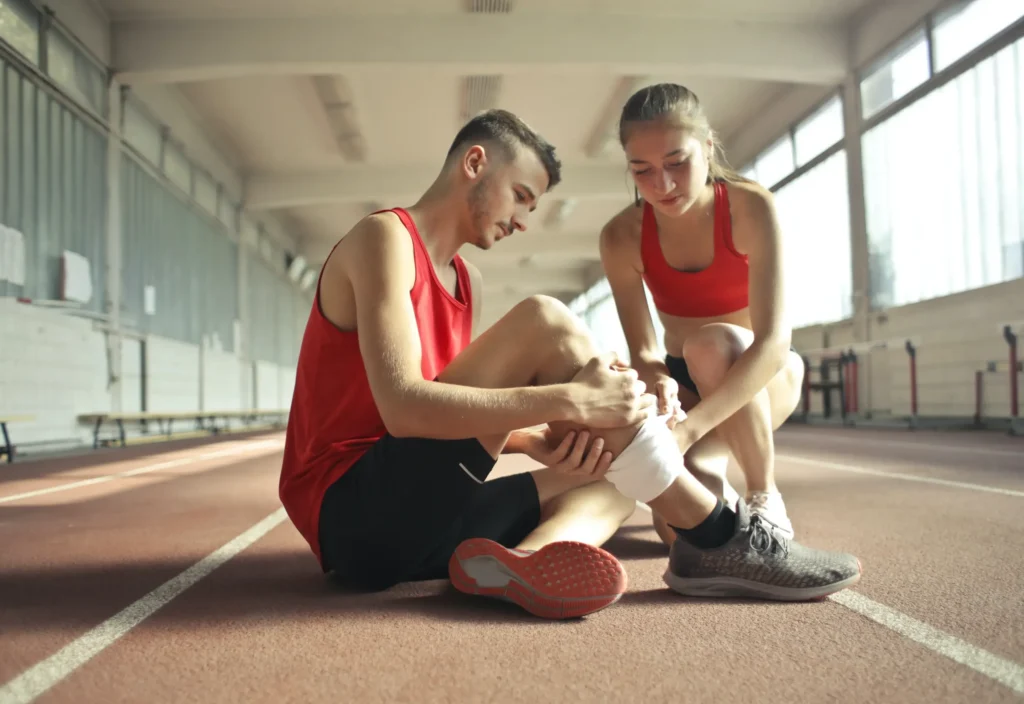 Injured man on the ground holding his leg while a woman wraps it, representing the need to seek medical treatment for pain after an accident.