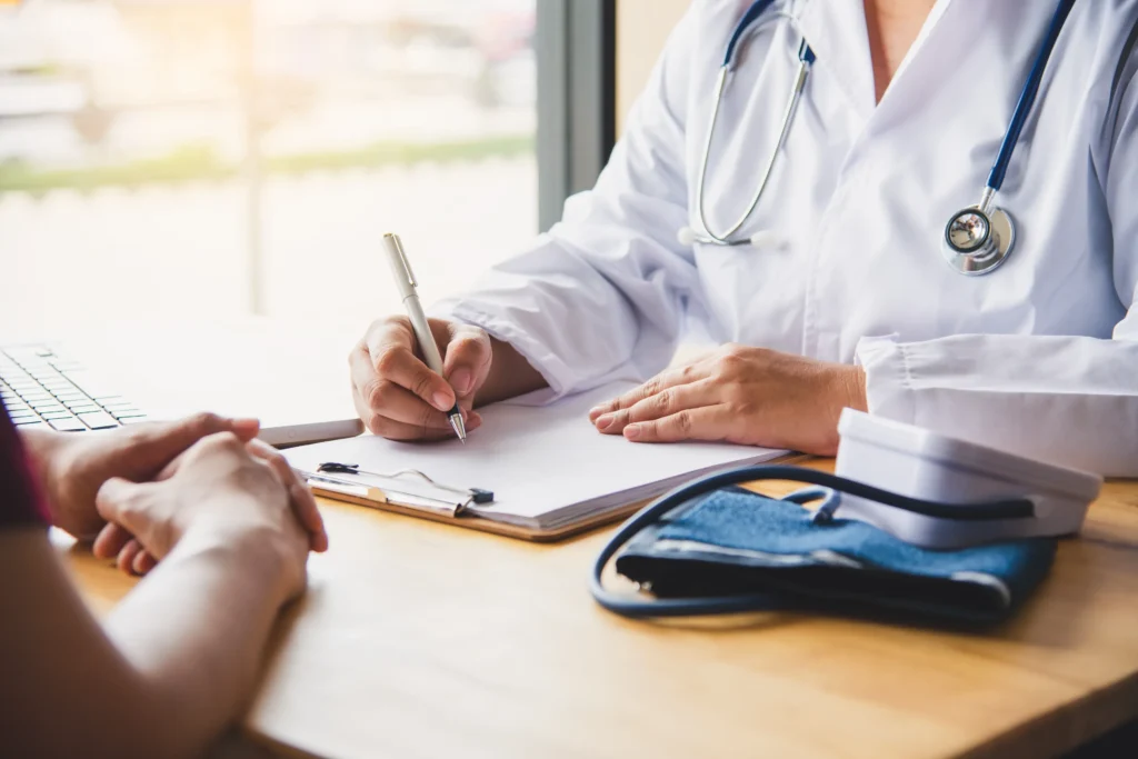 Close-up of a healthcare professional writing on a clipboard during a patient consultation, with a blood pressure cuff and stethoscope nearby.