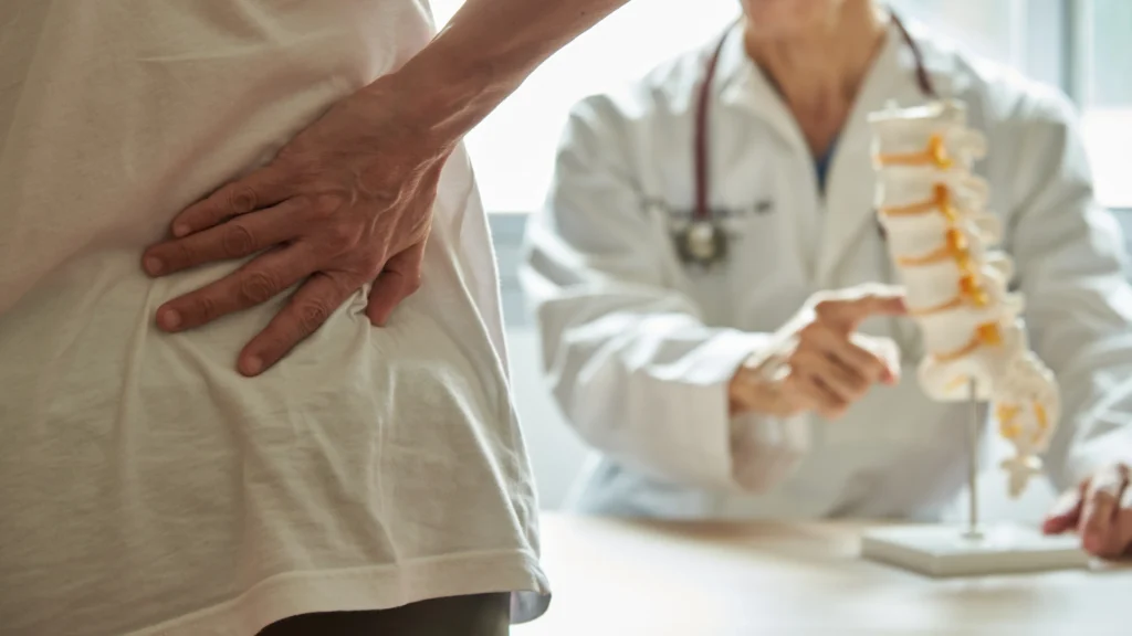 A patient with lower back pain consults a doctor, who points to a model of the spine, illustrating the medical evaluation process for chiropractic or physical therapy treatment options.