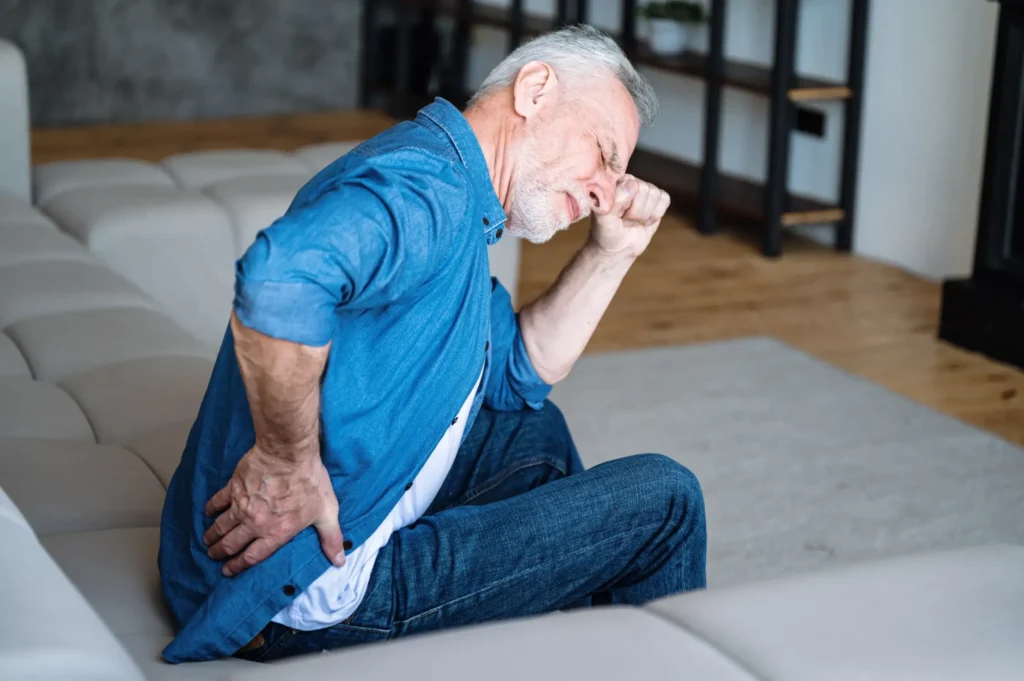 Older man sitting on a couch holding his lower back in discomfort, representing options for joint pain treatment.